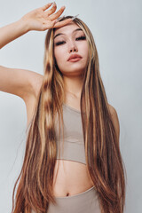 Fototapeta premium Young woman with long hair and natural makeup posing with hand above head wearing sleeveless top against plain background. Beauty, fashion and style portrait.