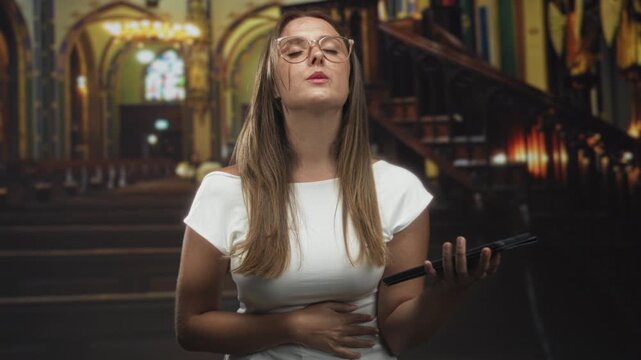 Woman holding tablet with one hand, other hand on stomach, eyes closed and head tilted up in church building; faith contemplation.