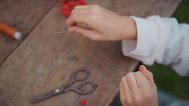 Closeup white hands threading red berries with thread and scissors on wooden table, delicate DIY garland making, cottage craft vibe, careful focused motion, tactile slow craft process, meditative