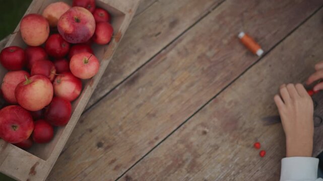Wooden table with apples and hands, crate of red apples beside spool of orange thread and scissors, child hands carefully threading small red berries into garland, rustic outdoor setting, cozy autumn
