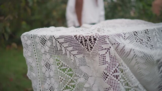 Outdoor rustic table being draped white woman and companion laying crocheted lace over weathered table in backyard hands smoothing fabric gentle laughter overcast sky seasonal greenery prepping