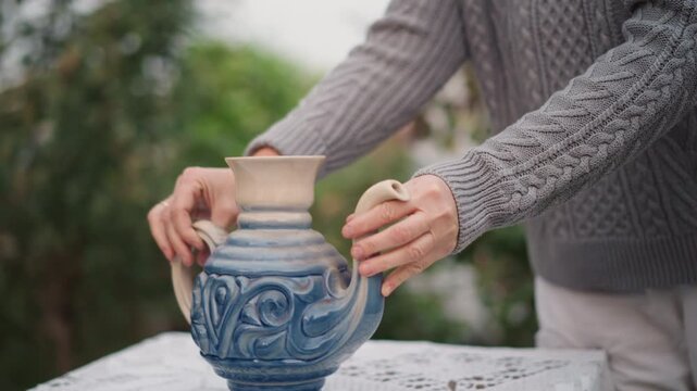 Hands placing ceramic blue jug on lace covered table outdoors person in gray knit sweater sets ornate indigo glazed teapot with decorative swirls, soft morning light, bokeh foliage background, careful