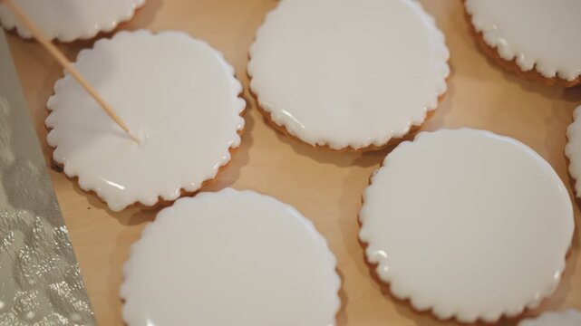 Closeup iced cookie with toothpick in quiet kitchen, slow careful decoration. Intimate frame shows hand using fine tool to shape glossy white glaze, subtle reflections, steady motion and focused