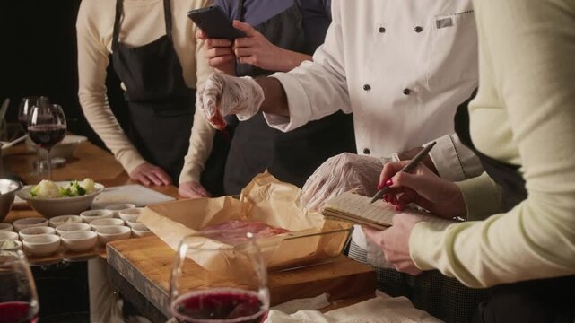 Tilt up shot of group of women taking notes on how professional chef seasoning and preapring meat for cooking during master class in restaurant