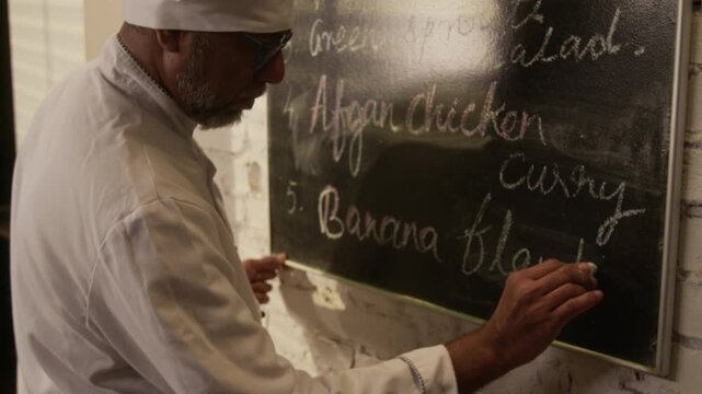 Low angle handheld zoom out shot of male chef in white uniform writing menu with chalk on black chalkboard in restaurant