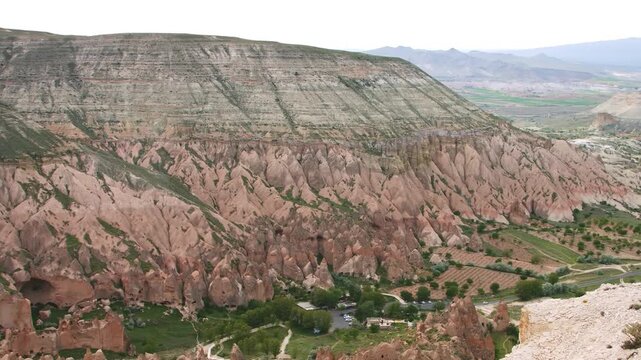 A wide, high-angle shot showcasing the unique, layered rock formations and vibrant pink/red hues of the Rose Valley in Cappadocia, Nevşehir Province, Turkey. The shot includes the dramatic slopes of t