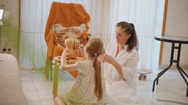White mother daughter decorating small pumpkin in bright living room with tiled floor and orange chair cover, mother braiding daughters hair while painting pumpkin, playful textures, scattered craft