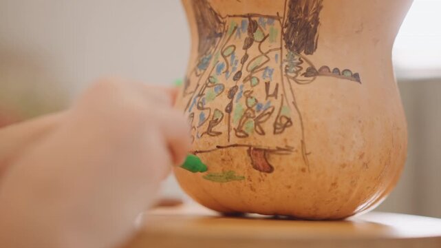 Young artist colors gourd on table. Child creates autumnthemed gourd artwork on wooden surface. Young child decorates pumpkin with green paint in cozy kitchen environment inspired by fall season