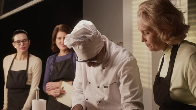Tilt up shot of biracial chef professionally cutting cucumber, while group of women watching him attentively during cooking master class in restaurant