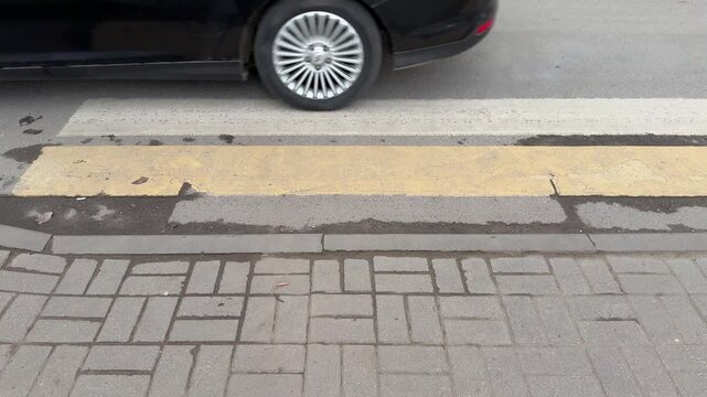 Low angle pedestrian crossing with passing car worn yellow white stripes, rough asphalt, concrete curb, patterned sidewalk, white.