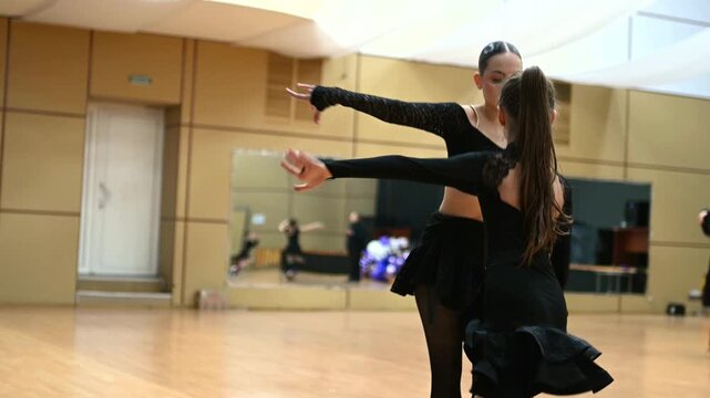 A young female dancer and a girl perform elegant choreography in a dance studio, likely in ballroom or Latin dance style.Both wear black performance outfits that highlight movement and body lines. 