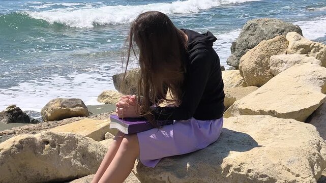 Young Christian Teen Girl Praying with Bible by the Rocky Seashore - Slow Motion Half Speed