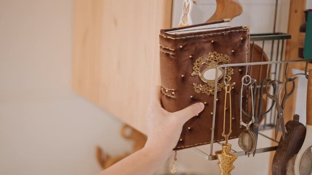 Hand retrieves ornate recipe journal near board kitchen, home baker pulls leather bound notebook from utensil rack with warm sunlight on cutting board, hints of spice and flour suggest pie