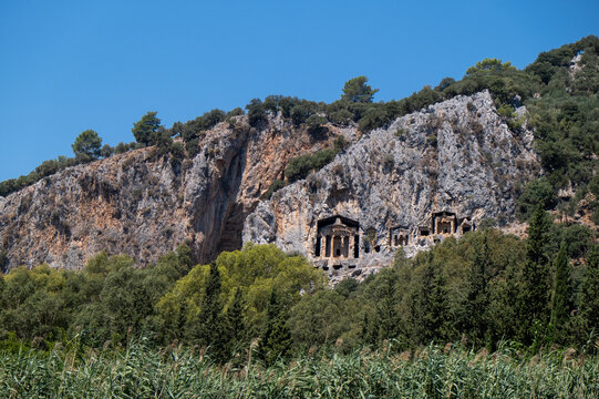 Ancient Lycian rock tombs carved into cliffs surrounded by forest landscape. Archaeological site, cultural heritage, historical architecture, nature and travel destination concept. Dalyan Ortaca Mugla
