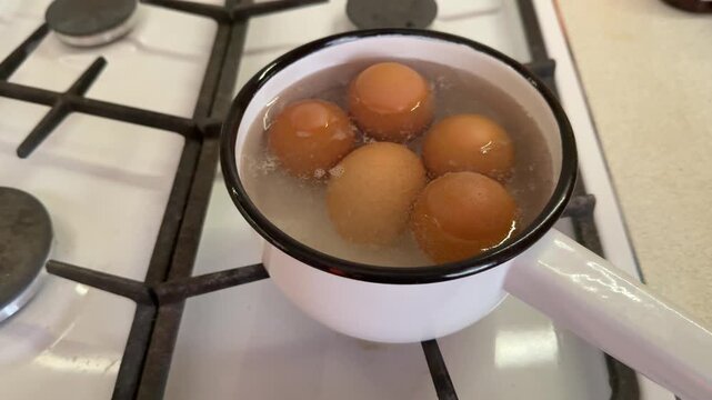 Close-up boiling brown eggs in ladle on gas stove, white enamel pot filled with bubbling water, gentle steam rising, homely morning.