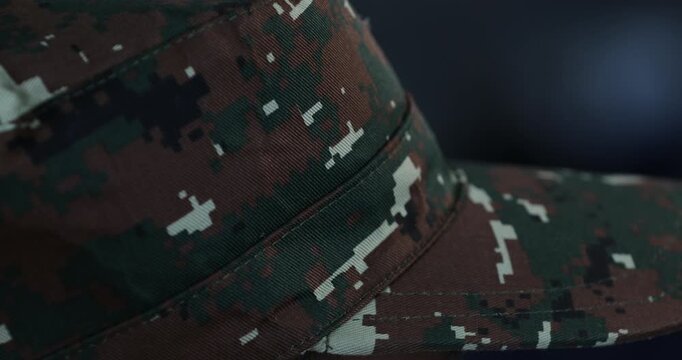 A close-up shot of a female hand gently adjusting the brim of a military camouflage cap. The pixelated camo pattern is in sharp focus against a dark background.