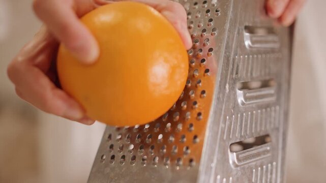 Closeup caucasian hands grating orange against stainless box grater, steady pressure producing fine zest, instructional kitchen tone, aroma release and culinary focus for garnish and baking
