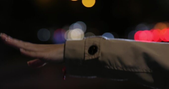 Close up shot of a woman's hand in a trench coat waving to hail a cab against a beautifully blurred background of colorful city traffic lights at night.

