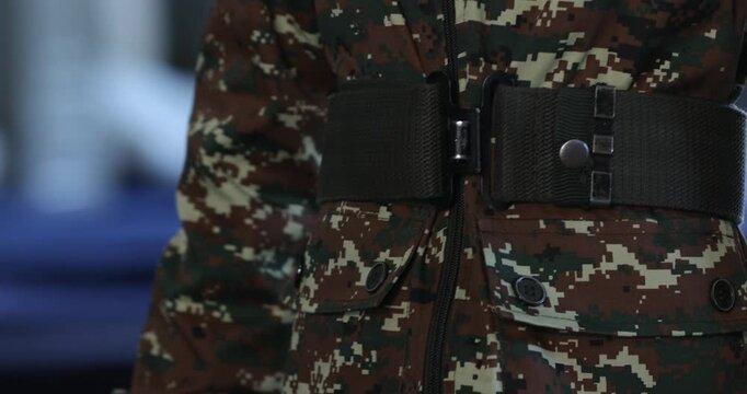 Close-up shot of an anonymous soldier in a digital camouflage uniform carefully fastening a wide, dark green tactical belt around their waist against a softly blurred indoor background.

