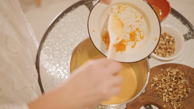 Overhead shot pouring orange zest into pumpkin pie batter, hands moving bowl toward stainless mixing pan, chopped pecans on wooden board, cozy holiday kitchen prep, casual baker arranging topping
