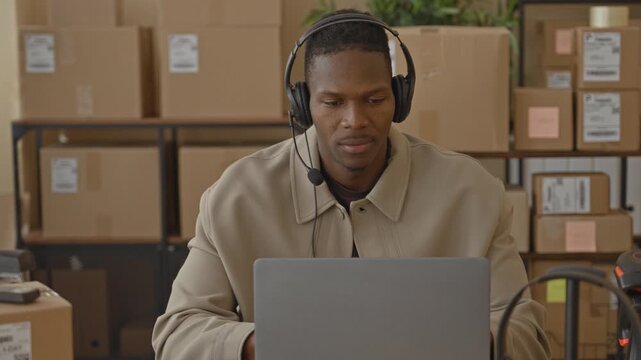 Man wearing headset and speaking into microphone while typing on laptop among parcels in building; customer support engaged.