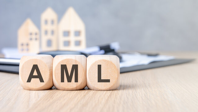 AML - Anti-Money Laundering - acronym on wooden blocks. Blurred background with miniature houses and magnifying glass. Real estate compliance concept.