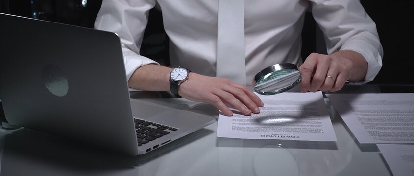 Businessman examining a document using a magnifying glass, focusing on fine print and terms in a legal agreement, working late. Audit and overworking concepts