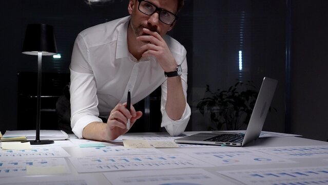 Dedicated businessman intensely analyzing financial documents and charts spread across his desk under a table lamp, concentrating on complex data in a dark office while working late evenings