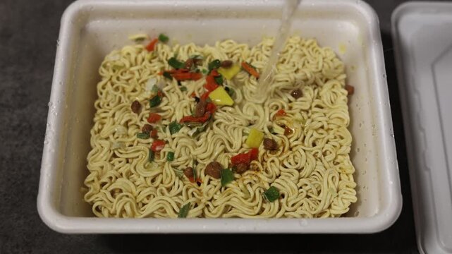 Close up video. 60 fps. Top view. Cook pours hot boiling water from kettle, over quick cooking instant egg curly noodles, ramen with spices, herbs in plastic plate for Asian soup. Healthy fast food