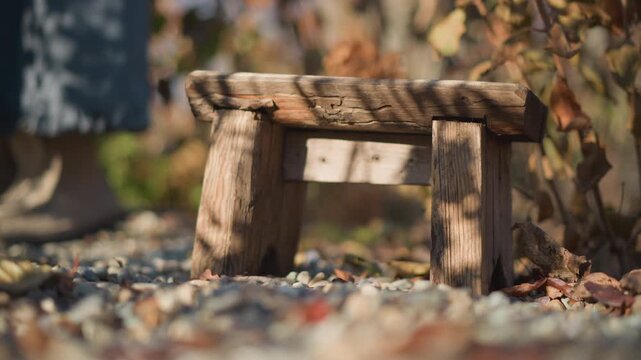 Rustic wooden stool on gravel, home garden scene with autumn leaves, weathered wood grain, soft sunlight, shallow depth of field, small handmade step suggests cozy homestead and outdoor kitchen mood,