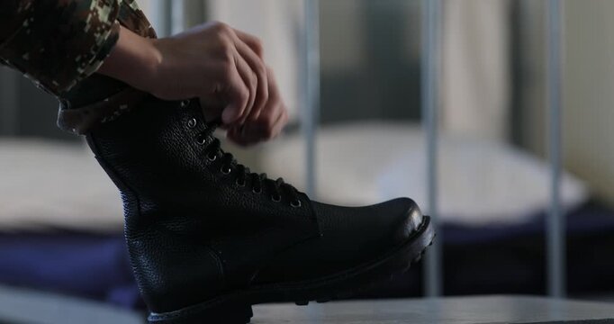 An unrecognizable soldier wearing camouflage pants is carefully tying the shoelaces of heavy black combat boots while resting one foot on a small stool in a barracks room.

