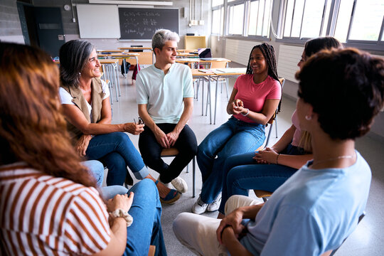 A diverse group of young adults and their instructor sit in a circle for an open dialogue session. They are smiling and sharing ideas in a spacious school room with a blackboard.