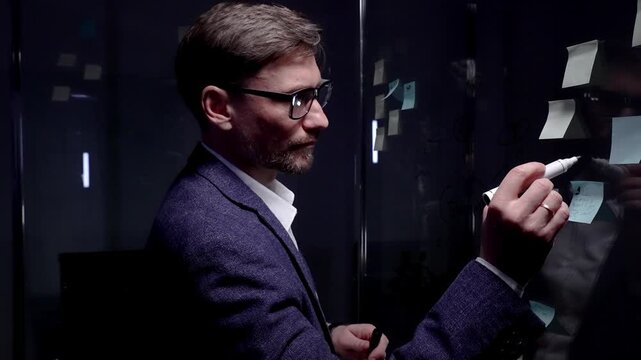Businessman planning new strategies, analyzing data on a digital tablet, and structuring ideas using sticky notes on a glass board in a modern office