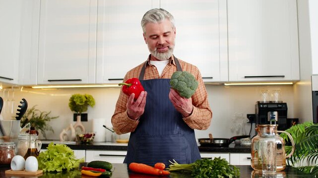 Middle-aged man smiling in home kitchen holding fresh pepper with broccoli for healthy diet choice and clean eating. Mature guy enjoying meal prep for weight loss nutrition plus fitness goals indoors.