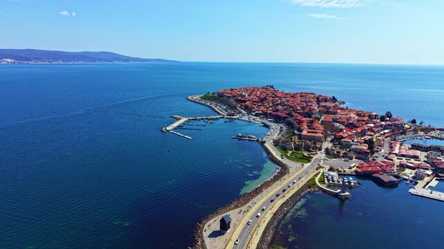 Nesebar Old Town shows narrow streets and red-roofed buildings by the water. Boats are docked in the marina with the sea in the background. Sunny weather highlights the landscape.