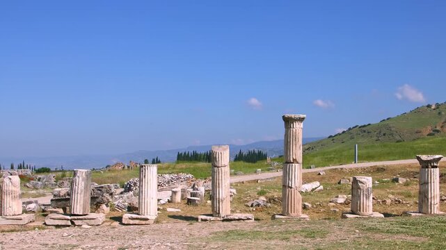 A low-angle, wide shot of several ancient marble columns standing among the archaeological ruins of the Roman city of Laodicea, near Denizli, Turkey. The site features a dirt ground, with green hills 