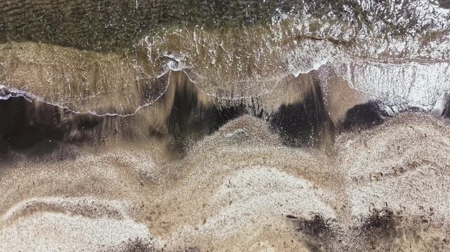 Waves gently roll onto the sandy shore as the tide goes out. Sand patterns mix with water in a coastal area during a sunny day. The scene captures nature's simple beauty.