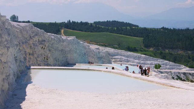 A wide shot of tourists walking and bathing in the stunning white travertine terraces of Pamukkale, Turkey. This UNESCO World Heritage site, also known as the "Cotton Castle," features natural hot spr