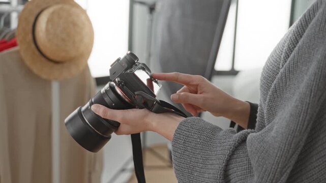 Woman photographer touches camera screen with finger, reviews settings and holds lens near a hat rack and window in studio; concentration.