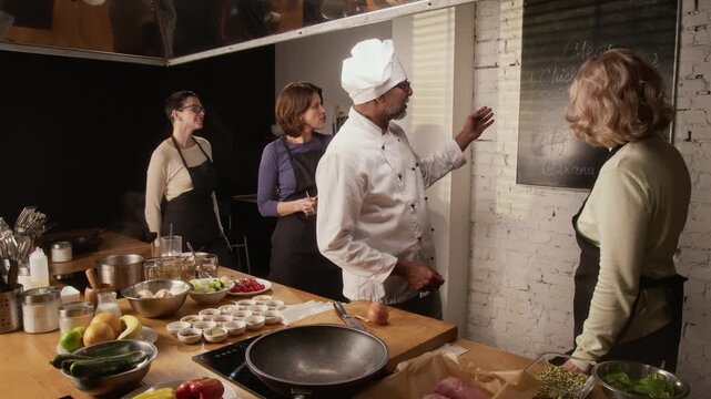 Middle aged biracial male chef introducing group of women, gathered for cooking master class, to plan on wall in restaurant
