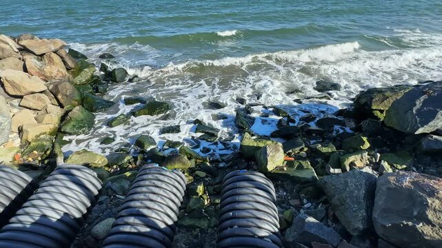 Waves crash against rocks by the shore while black pipes lay in a line. The scene captures the movement of water and the texture of rocks under sunlight.