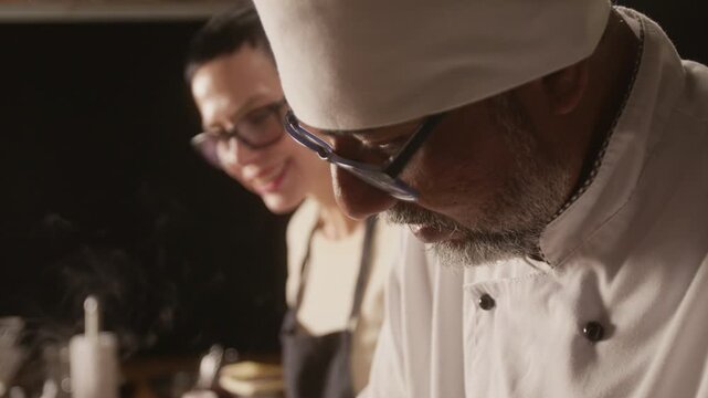 Close up tilt down shot of focused biracial chef in uniform putting salt into pan while cooking meat during group cooking master class