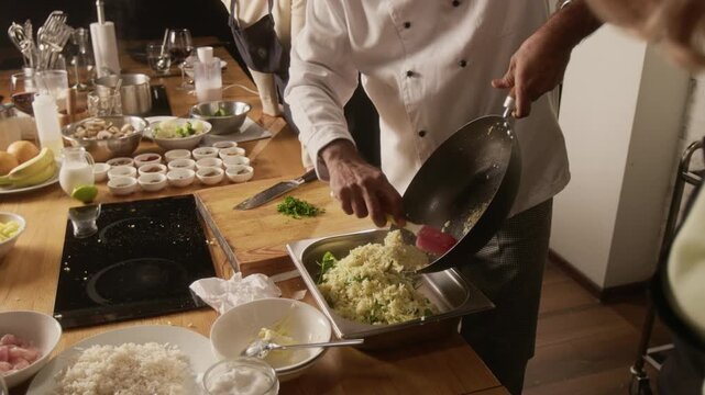 High angle tilt up shot of chef transferring rice from wok into container, surrounded by ingredients and participants, wearing aprons during cooking master class