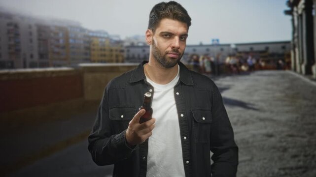 Man holding beer bottle, palm raised on a stone bridge with cobblestone walkway and city buildings; contemplation pause.