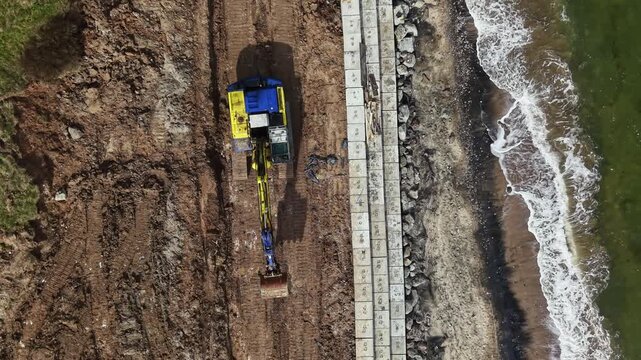 Heavy machinery digs into the earth as construction workers place concrete blocks for a seawall. Waves crash on the nearby shore while work continues.