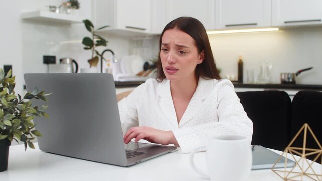 Young woman at home reads laptop result feels upset after lost game or rejected project clicks fast. Caucasian girl on sofa sits at table frowns stares at screen sad mood stress and worry deep sigh.