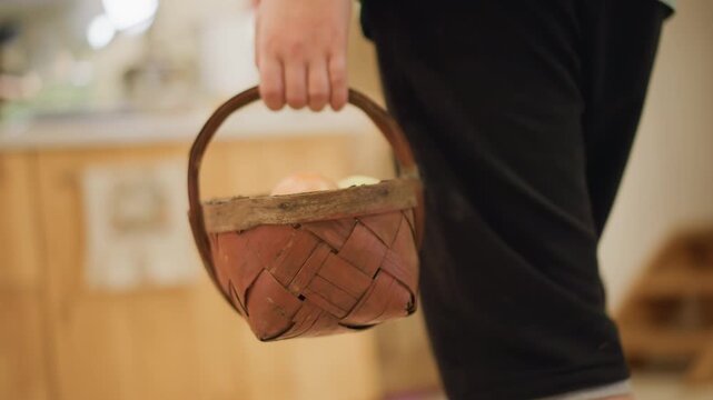 White teen carrying basket into kitchen. Young white helper walks from living room to rustic home cook space, holds woven basket of eggs and produce and places it on wooden counter. Ambient natural
