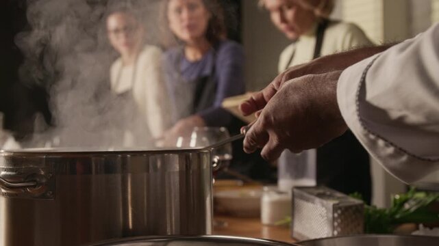 Low angle shot of unrecognizable chef cooking rice in pan while group of women discussing recipe during group cooking master class in restaurant
