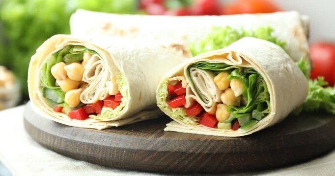 Board with pieces of tasty wrap with chickpeas, lettuce and vegetables turning against blurred background, closeup