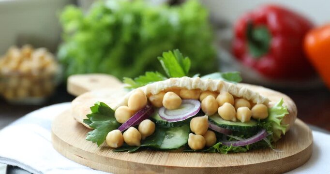 Board with piece of tasty pita bread with chickpeas, parsley and vegetables turning against blurred background, closeup